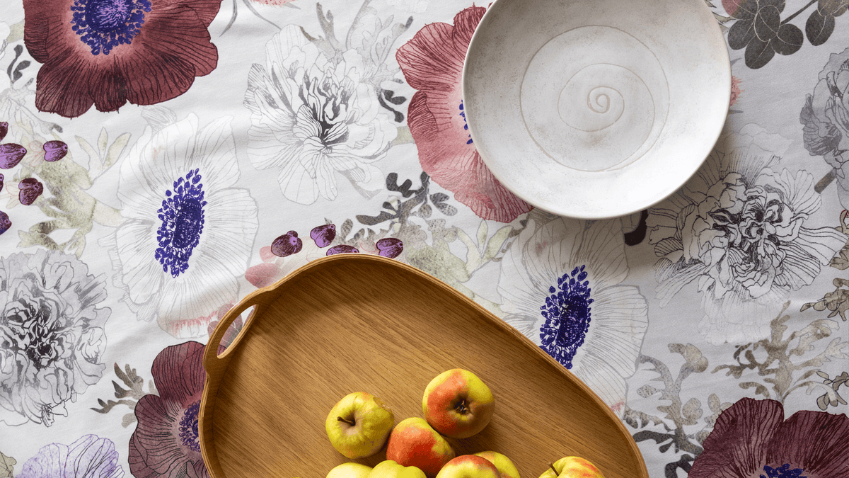 On top of the Leinikki tablecloth, depicted directly from above, a light-coloured Kivi series plate and a wooden Lautta tray with apples.