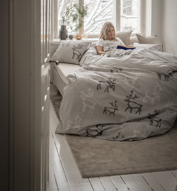 Woman reading a book on a bed with Saaga-patterned bedding in a bright room. Redirects to Shop the look collection.