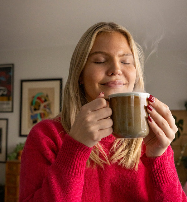 Woman in a red sweater holding a steaming Tuntu mug eyes closed. Redirects to Christmas table setting products collection.
