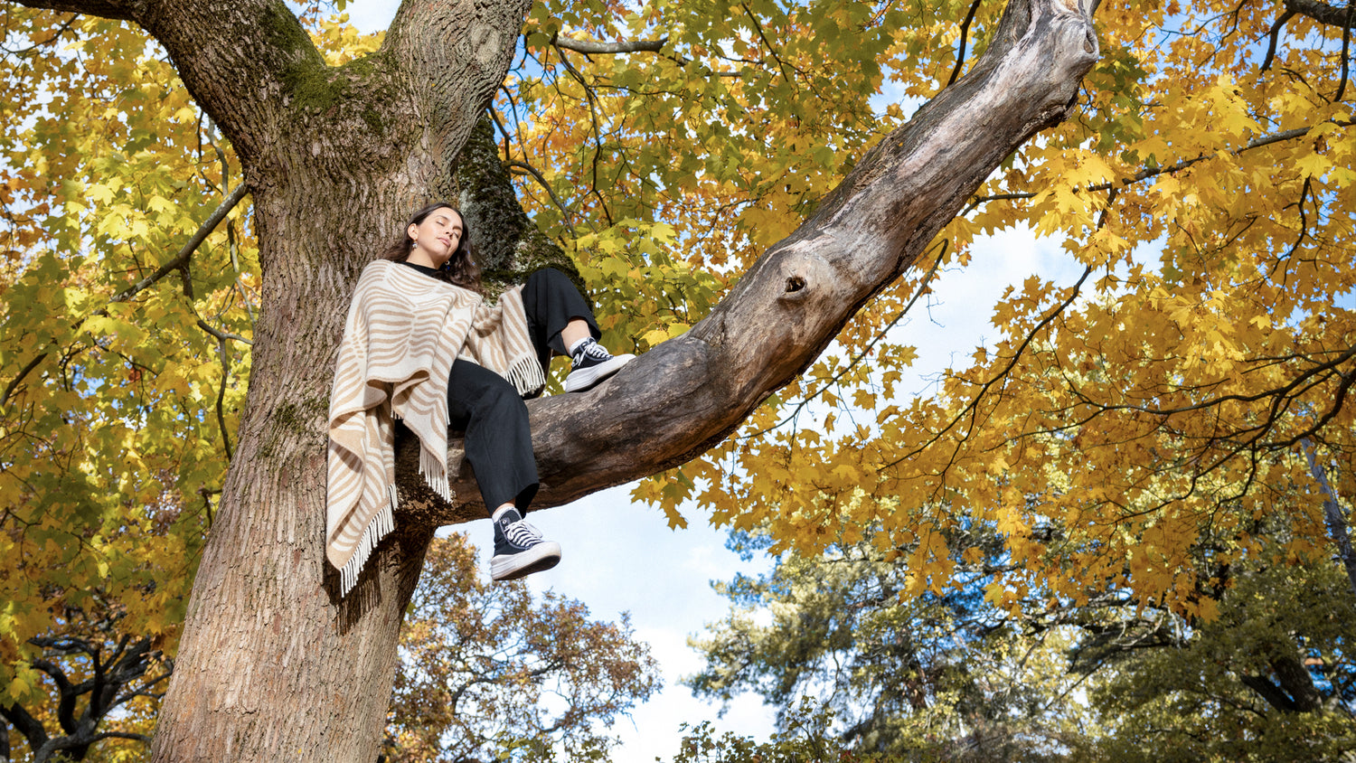 Young woman sitting on the tree branch eyes closed wearing a wool poncho from Vilja-collection.